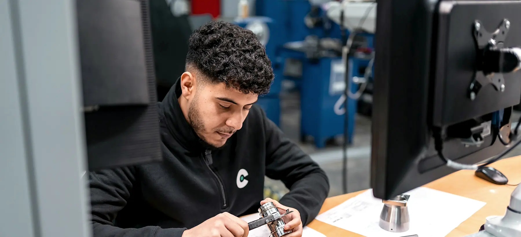A technician measuring a small machined component with a digital caliper at an inspection bench