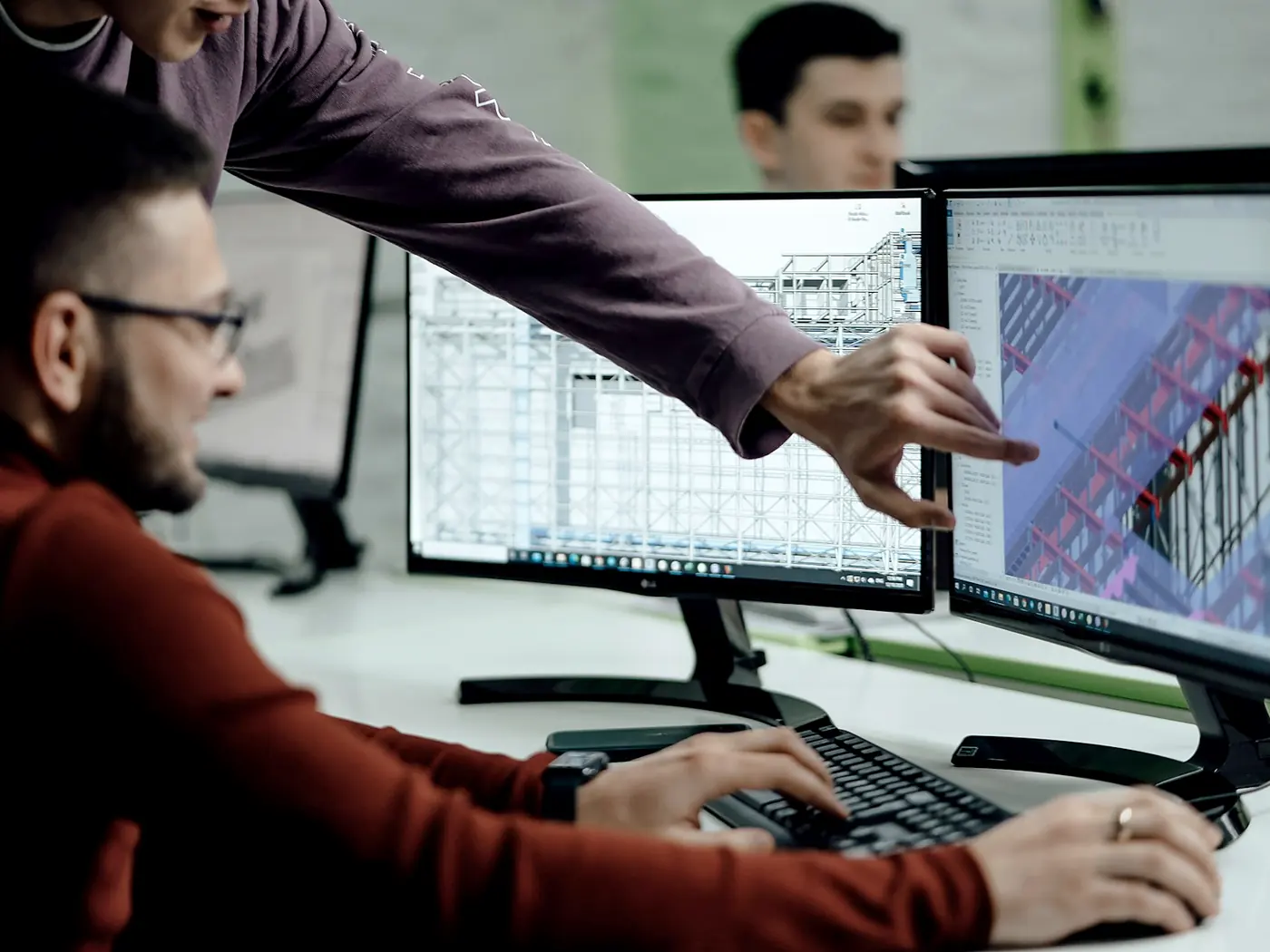 An engineer at dual monitors showing 3D CAD models, a colleague pointing at the screen
