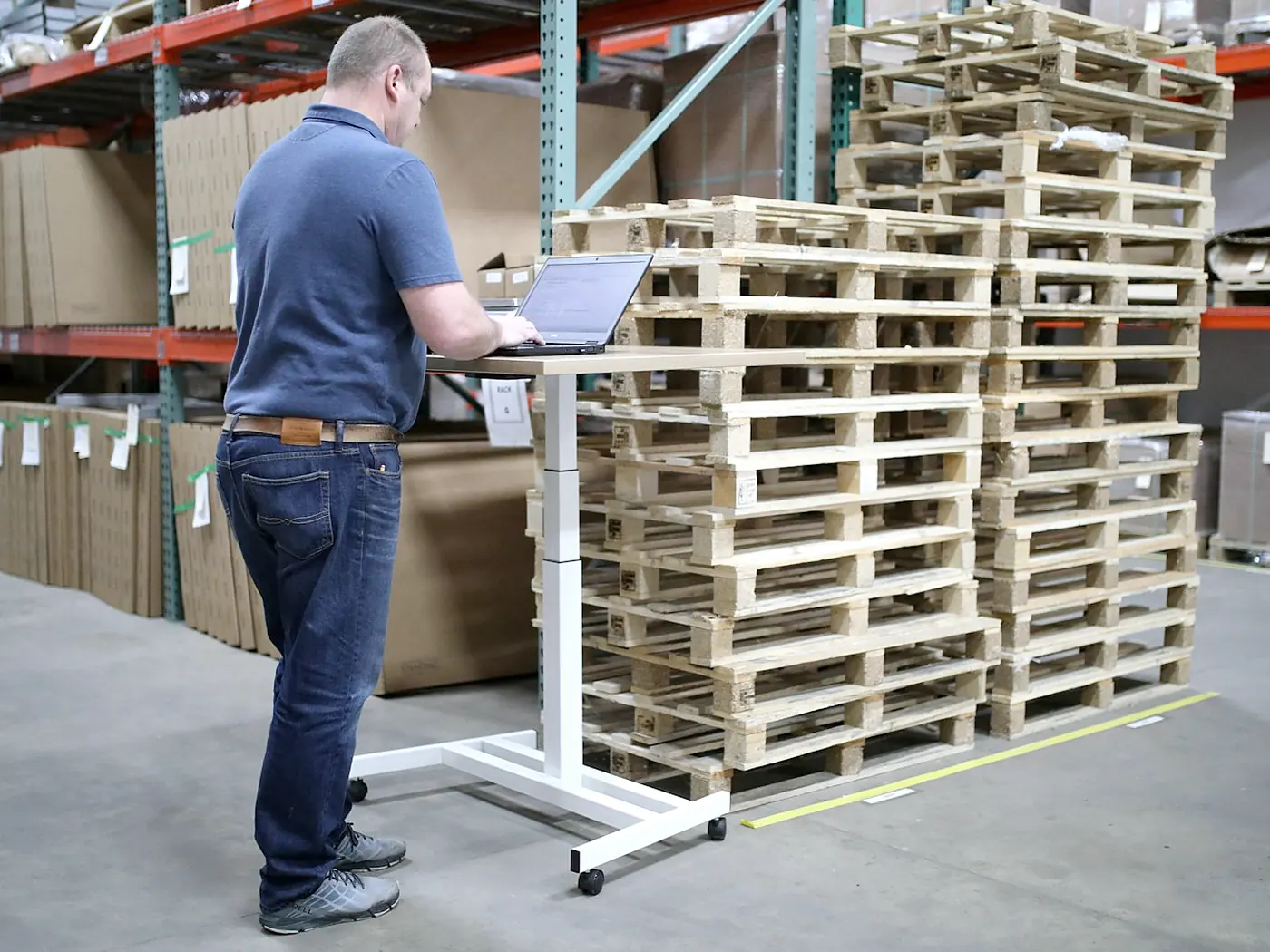 A worker at a laptop beside stacked wooden pallets in a warehouse